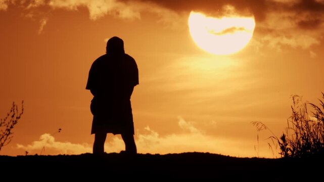 A silhouette of a man against the backdrop of an orange sunset and a big bright sun, the man is bouncing a basketball on the road