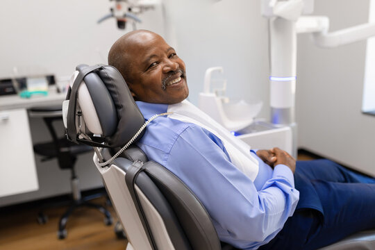 Smiling senior black male patient reclining in dental chair, looking back at camera beside dental x-ray equipment in modern clinic office, enjoying comfortable, positive dental care experience