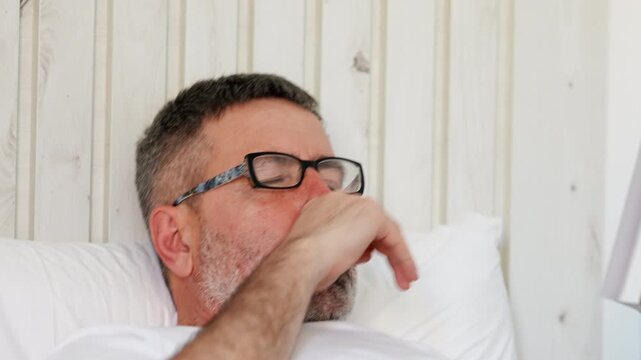Mature man with glasses yawning and falling asleep while reading a book in bed