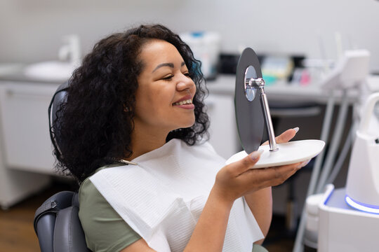 Happy black female patient smiling while checking her new smile in a handheld mirror after dental treatment, reflecting confidence, oral health and professional dental care
