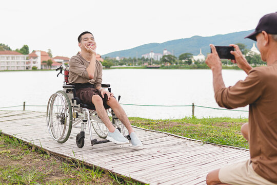 Happy disabled Asian young man on wheelchair posing for father taking picture with smartphone outdoors, inclusive family travel, accessibility lifestyle, caregiver, support and disability diversity