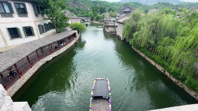 Boat with flower decor sailing on narrow river in ancient town. Traditional Chinese water village architecture with stone bridge. Tourism, travel and sightseeing in historic china city.