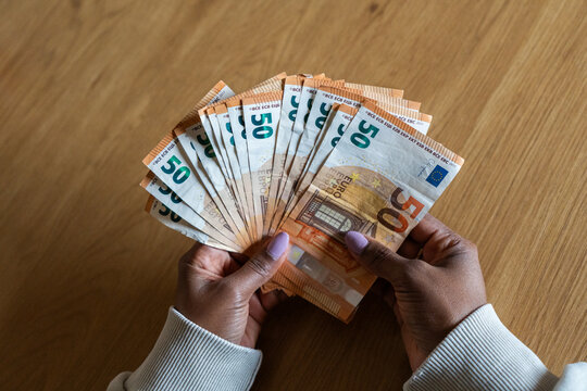 Close-up top view of a Black woman holding and counting a large amount of fifty euro banknotes over a wooden table, representing savings, income, wealth and financial planning.