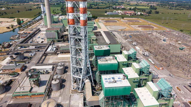 Aerial view of the Terna Tavazzano Est electrical substation with tall industrial chimneys, power infrastructure, and surrounding green fields, Montanaso Lombardo, Lombardia, Italy.