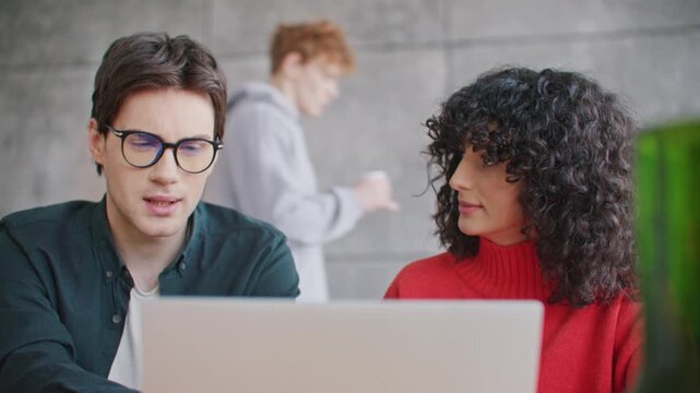 Young Caucasian man and woman discussing project while working together on laptop. Colleagues exchanging ideas in modern office. Team collaboration solving tasks and planning work.