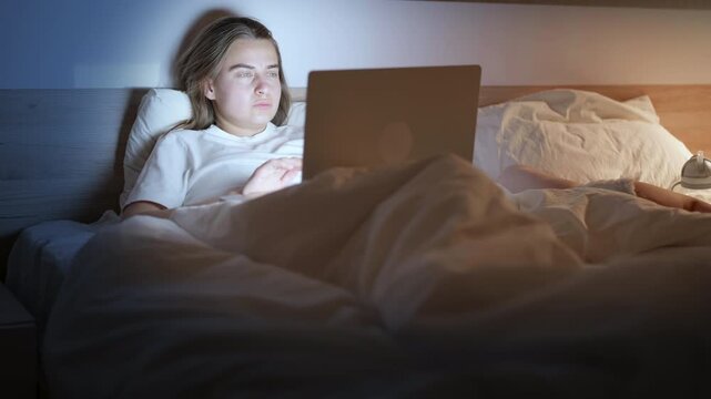 Young Caucasian woman sitting in bed at night, using a laptop, showing visible expressions of fatigue and frustration while wrapped in blankets