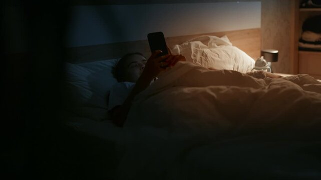 Young female using smartphone in bed, illuminated by soft light, surrounded by white bedding and wooden furniture in a cozy bedroom setting at night