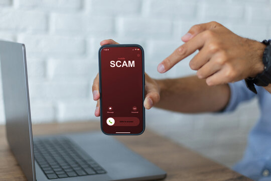 Man points to a smartphone showing a prominent SCAM call alert. Incoming call screen on a phone. Office desk with a laptop and a white brick wall background. scammers, concept