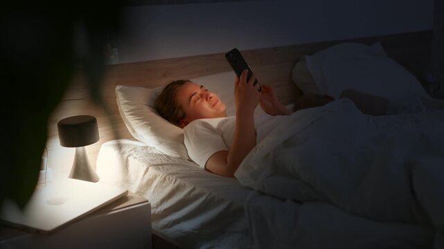Young woman in white shirt lying in bed using smartphone, illuminated by bedside lamp, cozy bedroom setting with soft bedding and warm lighting creating a relaxing atmosphere