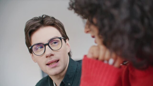 Young Caucasian man in glasses explaining ideas during teamwork meeting while smiling curly haired female colleague listening nearby. Creative collaboration in modern office environment.