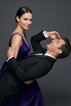 Elegant romantic couple posing in a studio. Confident beautiful brunette woman in a long stylish purple evening dress supports a happy laughing man in a classic black tuxedo leaning backward.