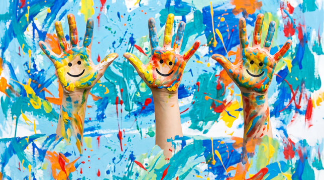 Painted hands with happy smiley faces raised high against a vibrant multicolored abstract paint background texture