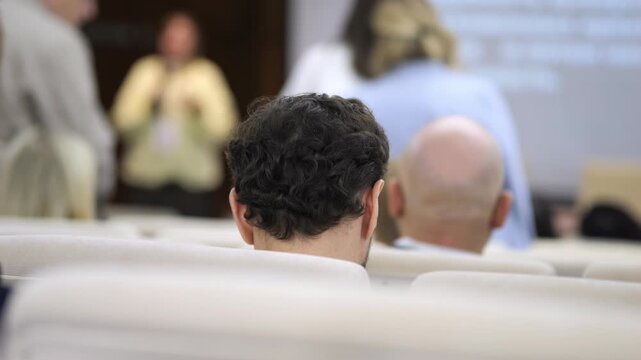 Audience seated in conference hall, focused attention. People watch presentation, heads slightly turned. Background shows blurred speakers and projector screen. Ideal for business meetings