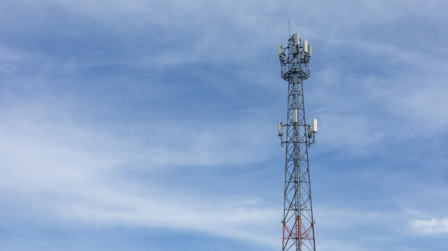 A cell tower rises high against a bright blue sky with scattered clouds. It is located in a rural area far from cities. The tower supports various communication signals