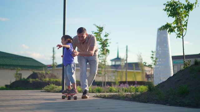 Father teaching little son to ride skateboard outdoors. Happy child balancing on board with dad support. Parent helps kid skate along park path. Family spends active time together in sunny city park.