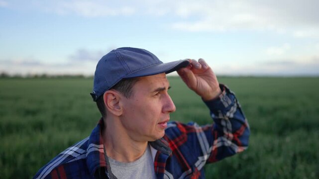 Thoughtful farmer in cap and plaid shirt stands in green wheat field. Man looks across crops and shields eyes with hand. Agriculture worker views farmland under sky. Farmer inspects growing field.