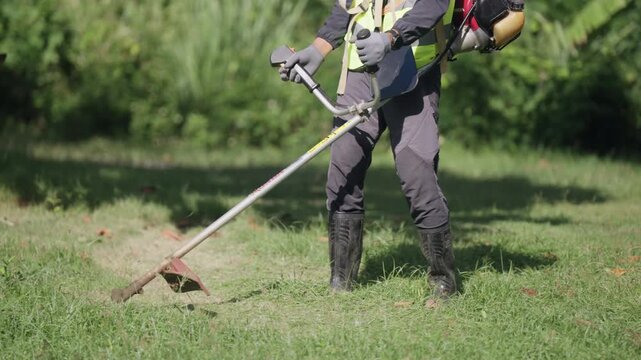 Grounds maintenance worker uses a brush cutter to trim grass in a green outdoor area while wearing protective equipment, representing landscaping, safety and property care.
