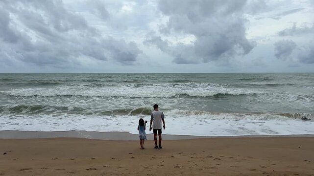 A child and an adult stand together on the beach, facing the ocean waves under a cloudy sky, enjoying a peaceful moment in nature.