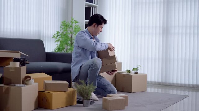 Young Asian man organizing and stacking cardboard boxes during home relocation in modern living room