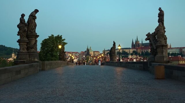 Historic Charles Bridge features cobblestone pavement, statues, and lanterns at dawn in Prague. Tourists walk toward Prague Castle. Czechia landmark represents European travel and heritage.