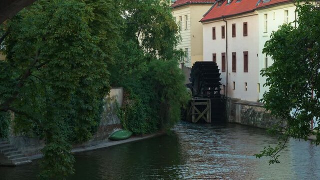 A historic wooden water wheel rotates on the Certovka canal in Prague. Traditional buildings line the urban waterway in Czechia. This scenic landmark illustrates European history and travel.
