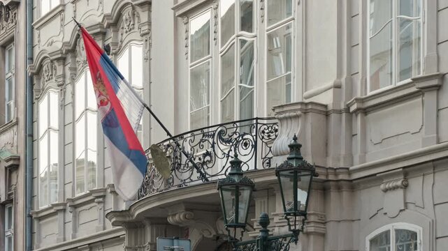 The national flag of Serbia flutters on a balcony pole of a historic embassy building. Classic architecture with lanterns represents government diplomacy, international relations, and politics.