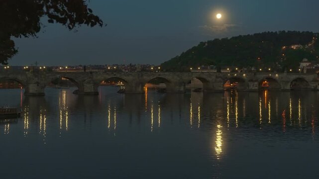 Historic Charles Bridge in Prague spans the Vltava River under a full moon at night. Golden illuminated lights reflect on calm river water. Ancient stone arch architecture shows European history.