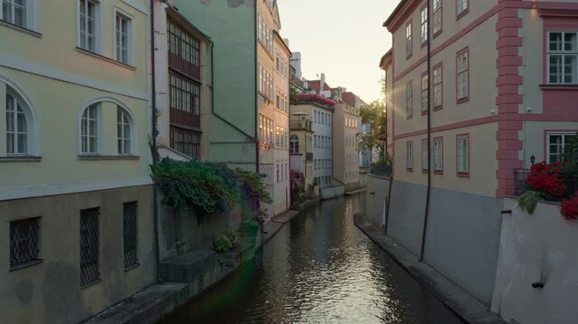 Historic Certovka canal flows between colorful old buildings in Prague. Warm morning sunlight shines through ancient stone facades along the European urban waterway. Scenic tourism view.