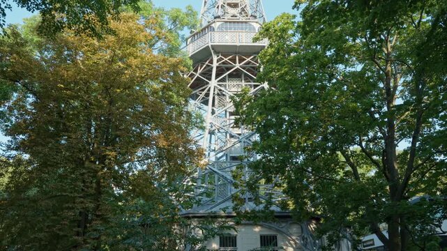 Petrin Lookout Tower stands among green park trees in Prague, Czechia. Steel lattice observation tower rises into clear sky. Historic landmark represents European travel and sightseeing.