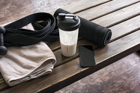 Gym equipment laid out on a wooden surface, including a shaker bottle, yoga mat, dumbbell, resistance band, towel, and smartphone. Ready for a workout