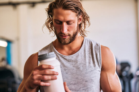 A man in a gym holding a protein shake. He is wearing a grey tank top and has a beard. The background is blurry, but appears to be a gym with exercise equipment.