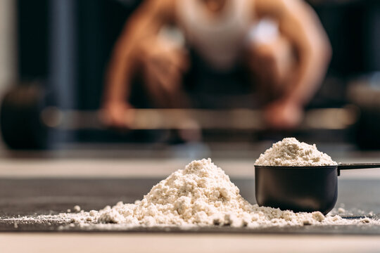 A scoop of protein powder and a pile of powder on a mat with a blurred background of a person lifting weights