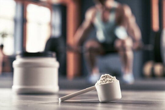 Scoop of protein powder beside a container in a gym with a blurred background of a man working out