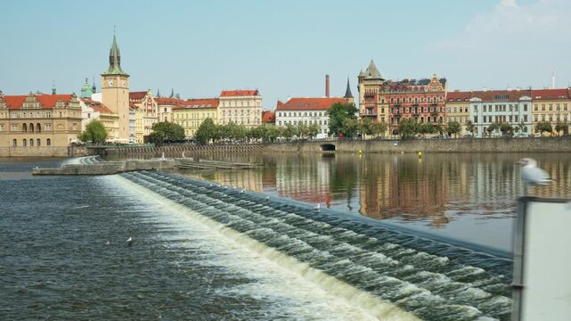 A river weir cascades water on the Vltava River in Prague during daylight. Historic Old Town buildings and water tower line the embankment. Scenic Czechia landscape offers travel destination.