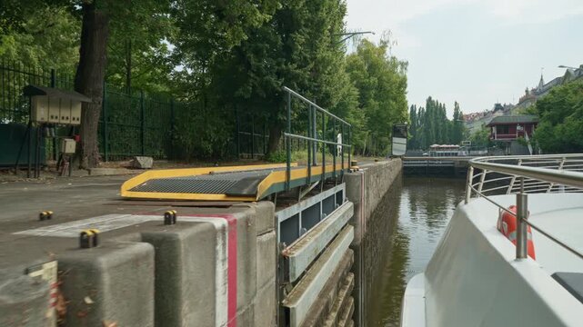 A tourist cruise boat sails past Smichov Lock stone wall in Prague, Czechia. Yellow mooring bollards and bridge structures line the waterway. Hydraulic engineering travel background scene.