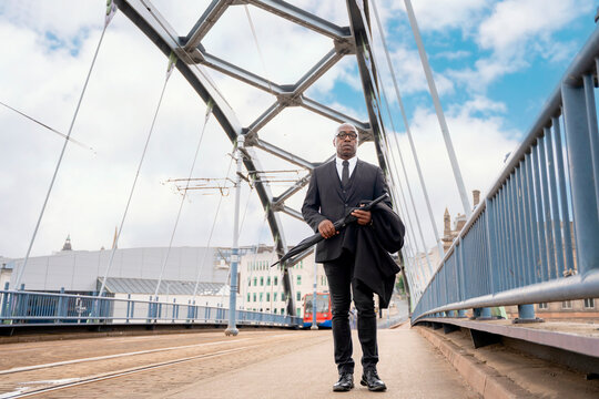 Businessman walks on a bridge during the day while holding an umbrella in hand and wearing a suit