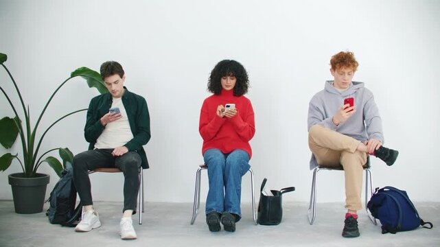 Three young people sitting on chairs and using smartphones in bright waiting room. Diverse friends ignoring each other while scrolling social media. Modern digital lifestyle with copy space.