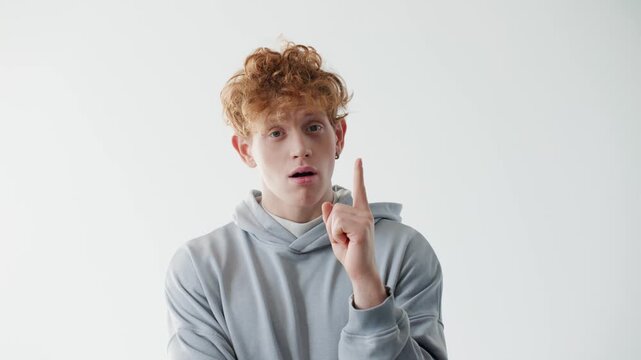 Confident young Caucasian redhead man standing with folded arms and rising finger like having idea in bright studio. Casual male wearing gray hoodie and showing calm self assured expression.