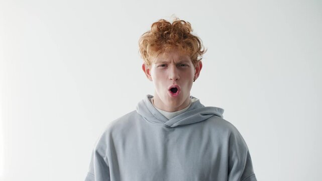 Excited young Caucasian man celebrating success with raised fists and playful expression. Redhead male wearing casual hoodie and cheering in bright studio. Victory emotion and positive energy.