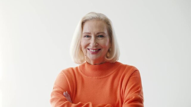 Confident senior Caucasian woman standing in orange sweater and looking at camera with calm expression. Elegant elderly female posing in bright studio. Self assured attitude with copy space.