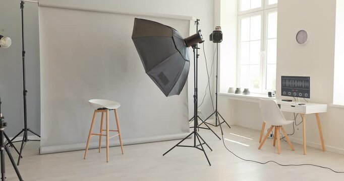 Studio lighting backdrop equipment umbrella stool workspace. Modern studio with grey seamless, softbox, computer desk by window and stool for portraits. Clear modern studio setup concept.