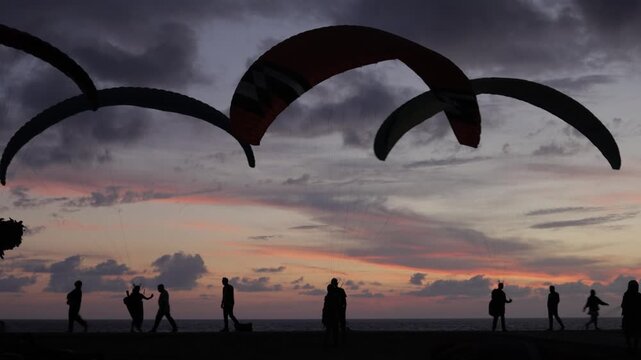 Silhouettes of paragliders and people against a vibrant sunset sky over the sea.