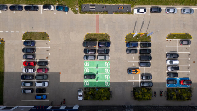 Aerial view of a parking lot featuring designated electric vehicle charging spaces marked in green, surrounded by various parked cars and landscaping in Gdansk, Poland.