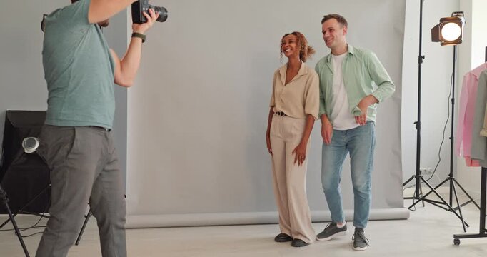 Photographer guides couple models in studio photoshoot. Professional portrait setup with soft lighting on backdrop. The pair pose with natural smile for camera. Team trust and service concept.