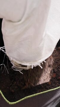 Vertical video. Woman Pours Dark Soil from a White Bag into a Large Black Fabric Pot, Preparing Fertile Substrate for Container Gardening on a Wooden Terrace