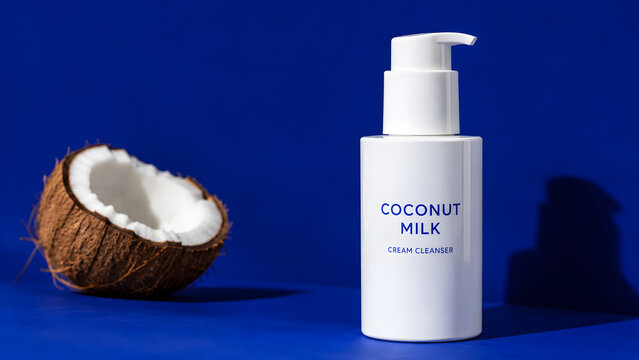 Coconut milk cream cleanser in a white pump bottle next to a half coconut on a blue background