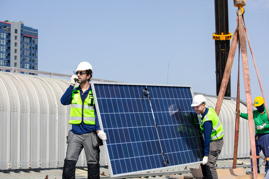 Solar technicians in hard hats and reflective vests guide a crane-lifted photovoltaic panel on an industrial rooftop, using radio communication for safe teamwork and renewable energy installation.