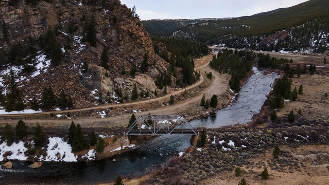Aerial view of a steel truss railroad bridge spanning a river, surrounded by rugged mountains, evergreen trees, and patches of snow in Buena Vista, Colorado, United States.