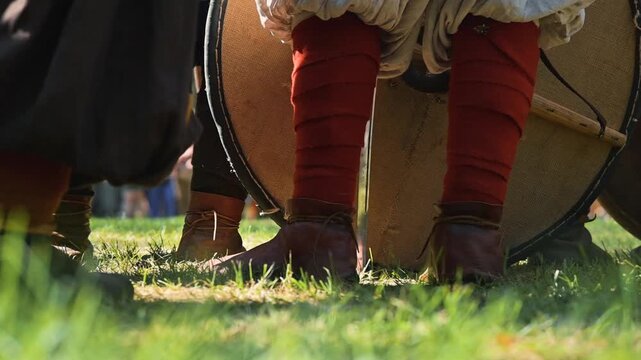 Medieval reenactors gather in a grassy field, preparing for the upcoming battle. Participants wear traditional clothing and hold drums, creating a lively atmosphere