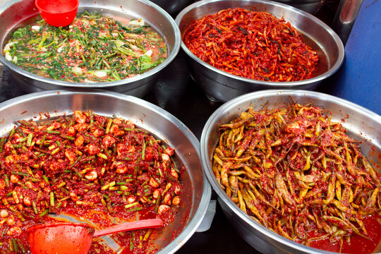 Large stainless steel bowls filled with various types of kimchi are displayed in a market setting, in Busan, South Korea.